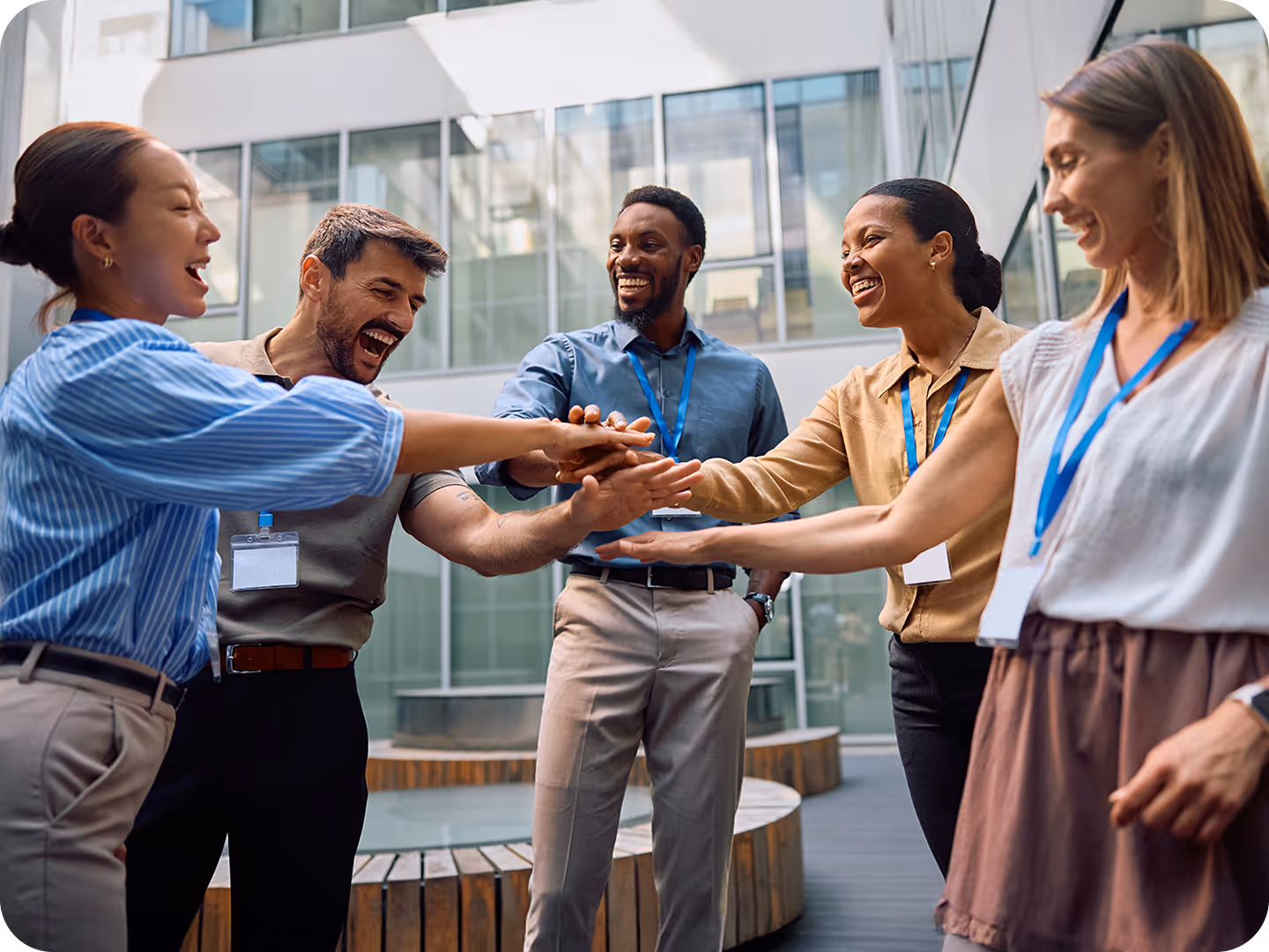 A diverse group of laughing coworkers huddling together with their hands in the center for a team-building exercise in a modern office courtyard.