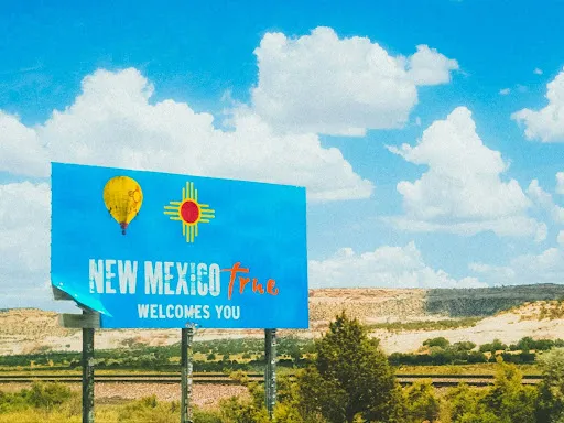 a sign along the New Mexico highway reads "New Mexico (True) Welcomes You" against a arid environment and a bright blue sky with clouds
