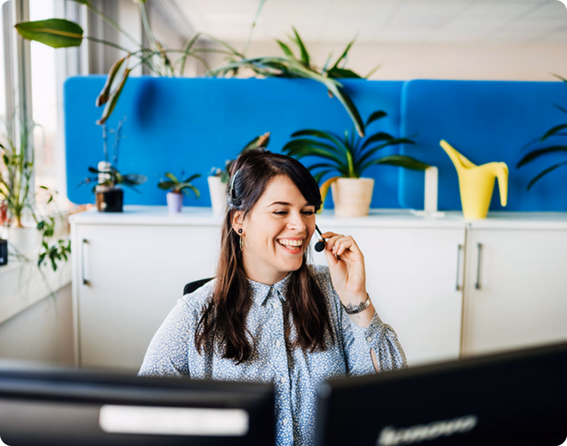 Smiling woman wearing a headset, sitting at a desk with computer monitors in a bright office with plants in the background.