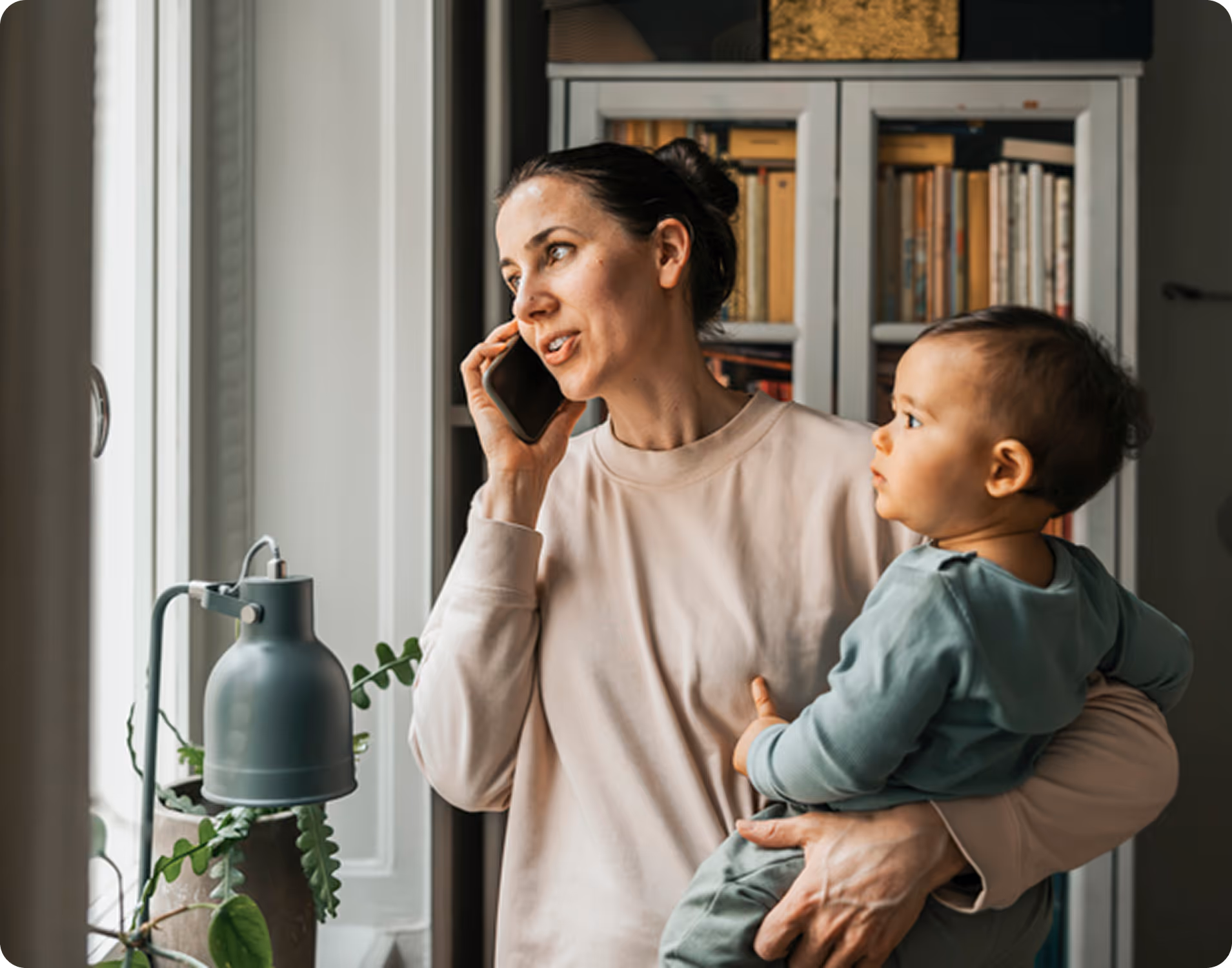 Woman holding a baby while talking on a mobile phone near a window with a lamp and plant nearby.