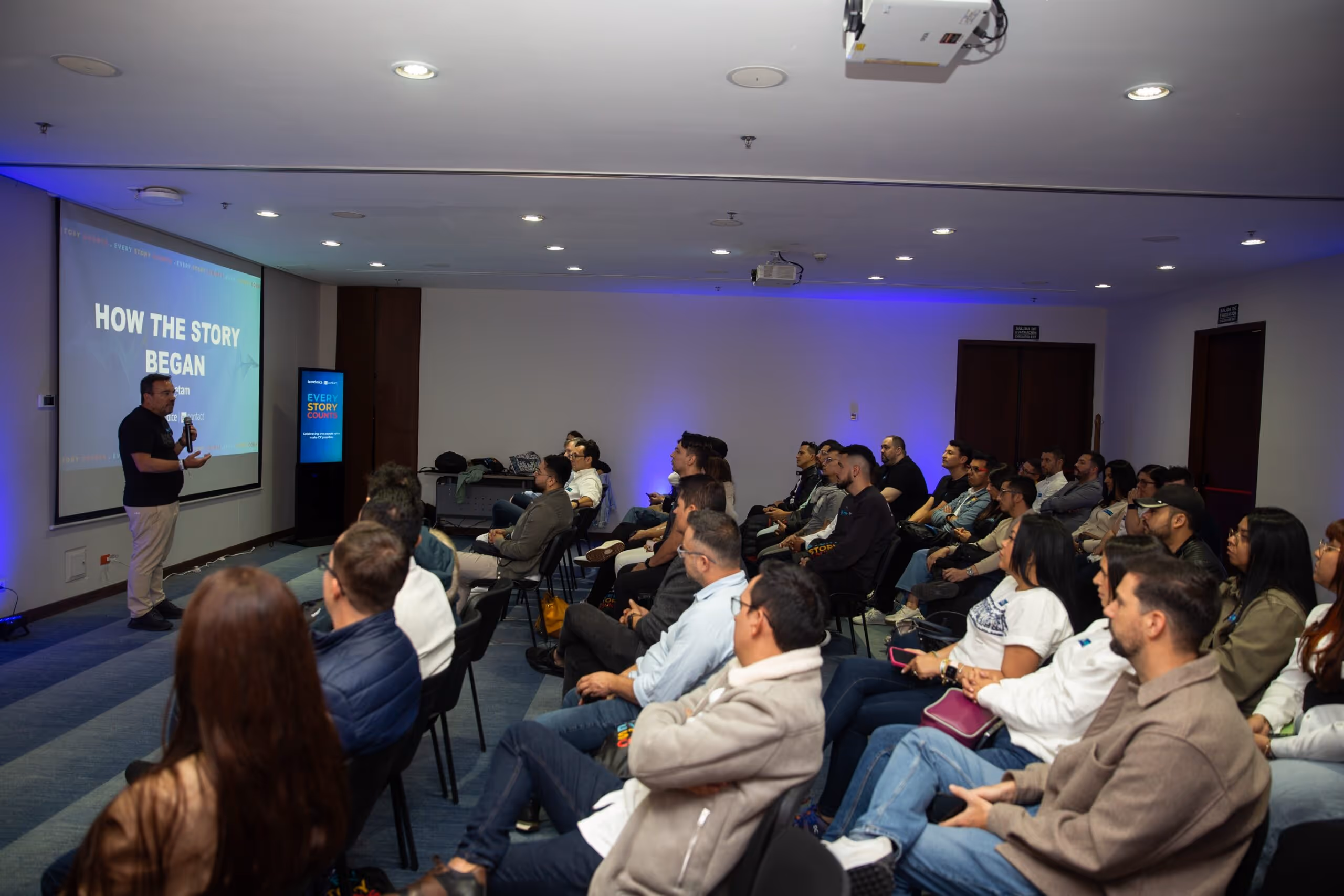 Man giving a presentation titled 'How the Story Began' to an audience seated in rows in a conference room.