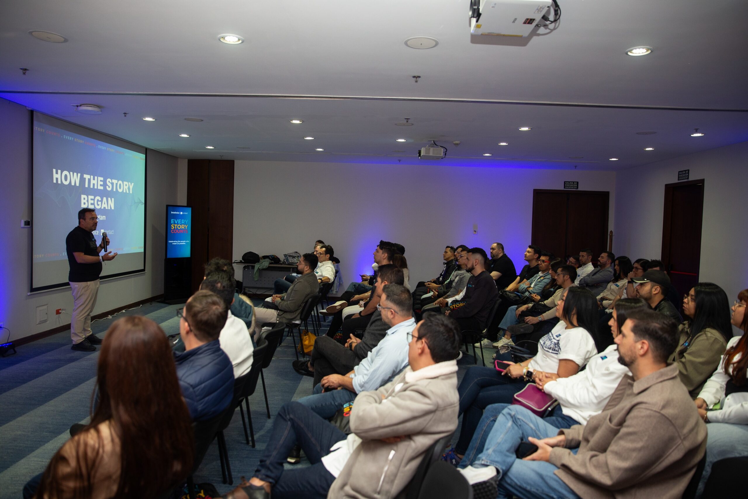 Man giving a presentation titled 'How the Story Began' to an audience seated in rows in a conference room.