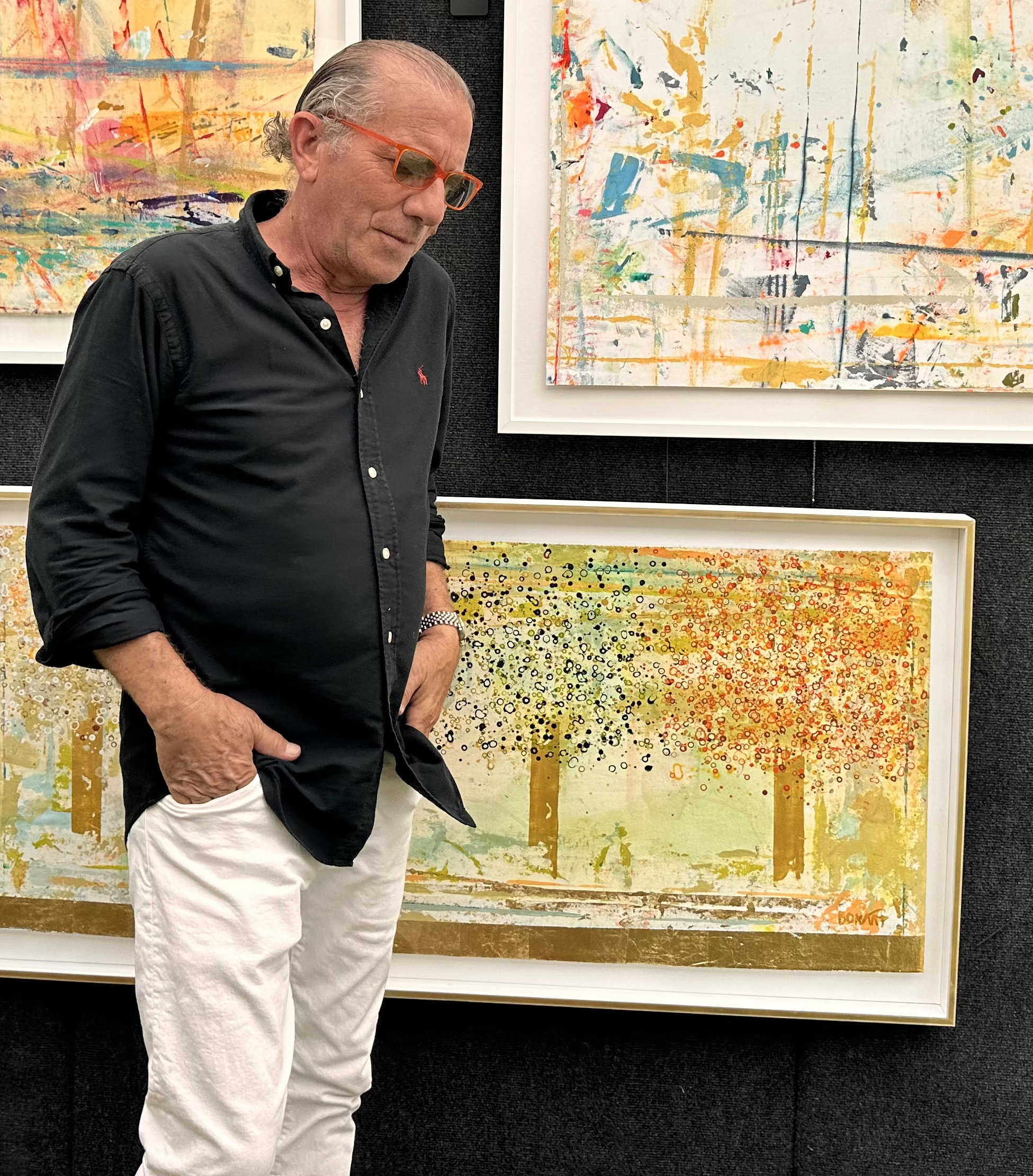 Ben stands in a minimalist living room with a brown leather and chrome chair, dark green side table, black stove in fireplace, and abstract yellow and with an abstract blue, green, and gold painting above a credenza.