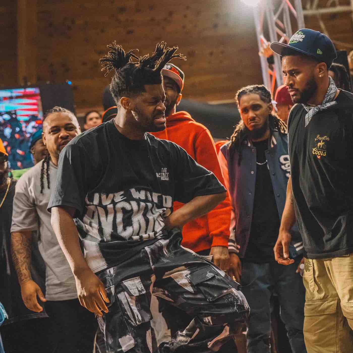 Man with styled dreadlocks wearing a graphic black t-shirt and patterned pants expressing intense emotion while a group of men watch in the background at an indoor event.
