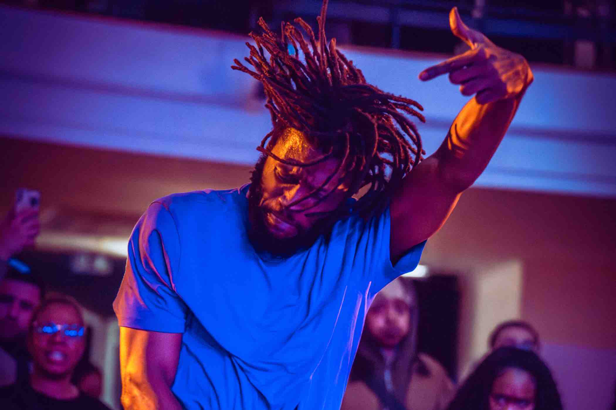 Man with dreadlocks dancing energetically in a blue shirt under colorful stage lighting with an audience in the background.