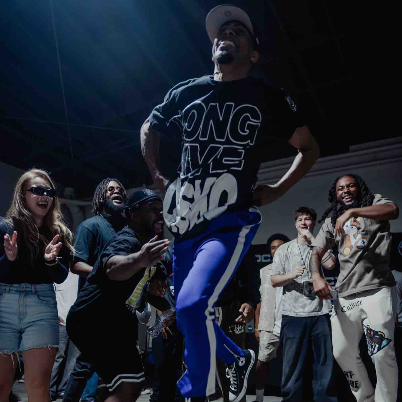 Man in blue pants and black shirt dancing energetically while a diverse group of people clap and cheer around him indoors.