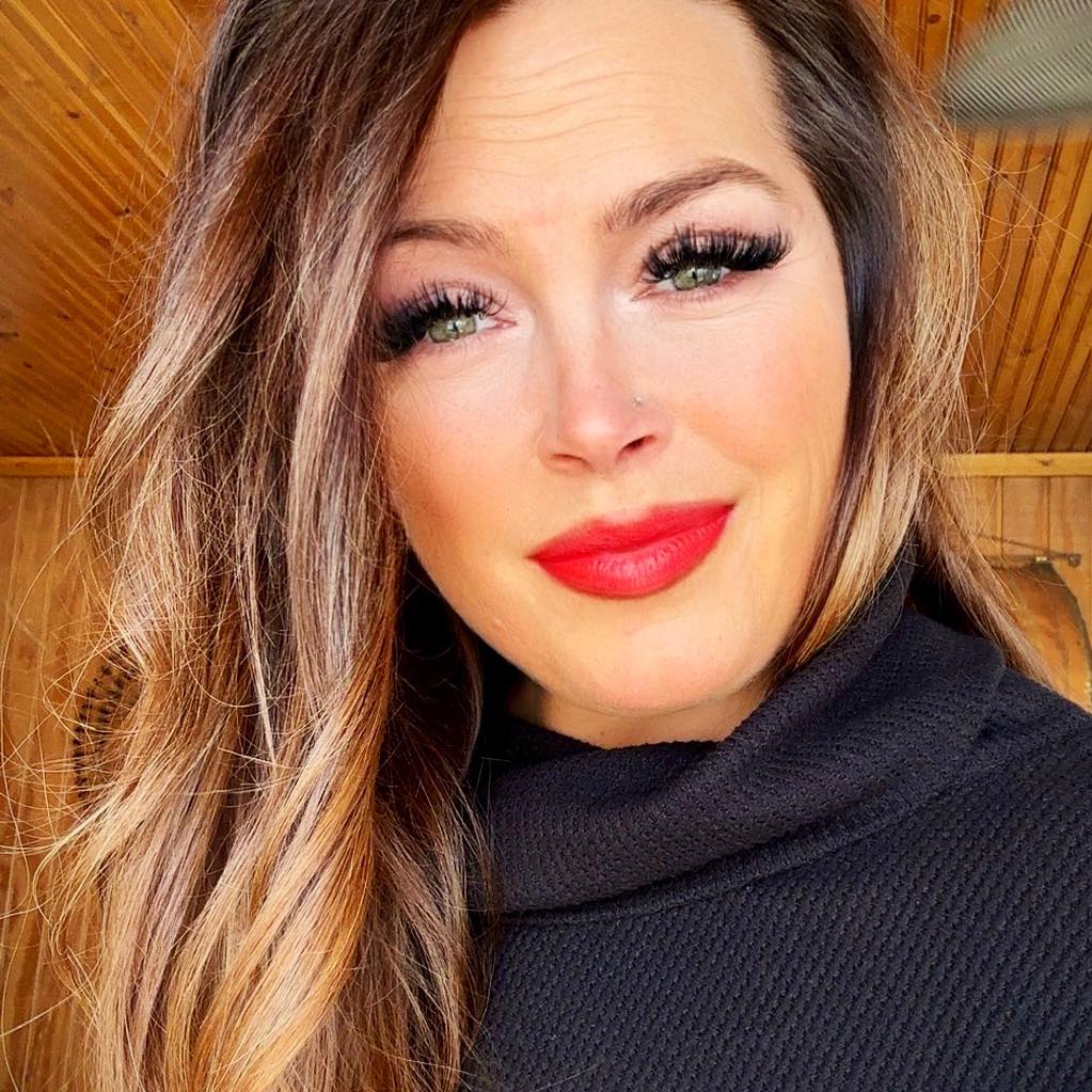 Close-up of a woman with long wavy hair, bold red lipstick, and long eyelashes, wearing a black top with a wooden ceiling background.