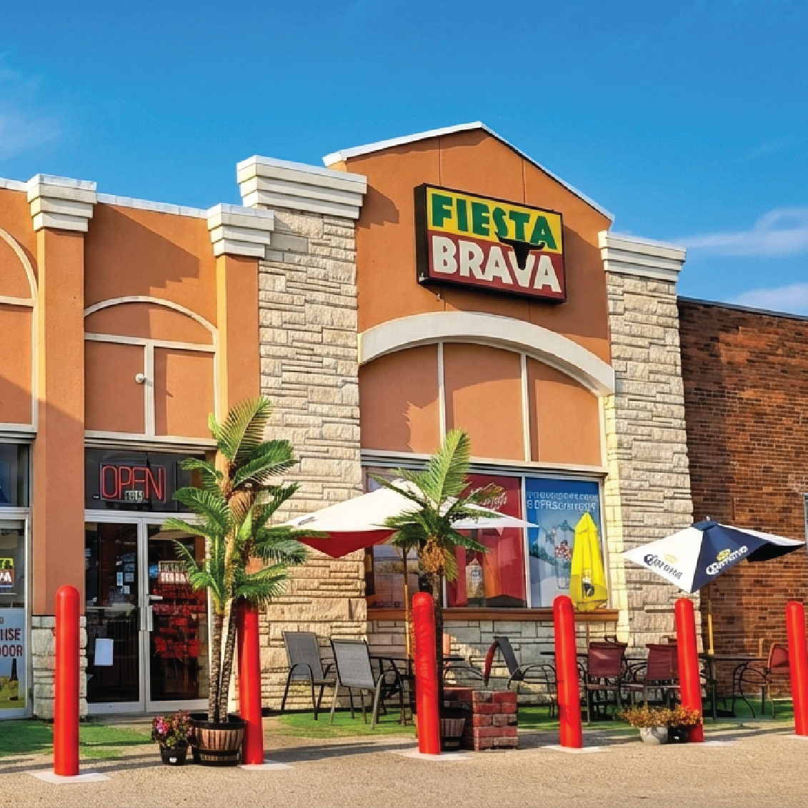 Exterior of Fiesta Brava restaurant with palm plants, outdoor seating, umbrellas, and red bollards under a clear blue sky.