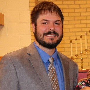 Man with dark hair and beard wearing a gray suit jacket, blue shirt, and striped tie, smiling indoors.
