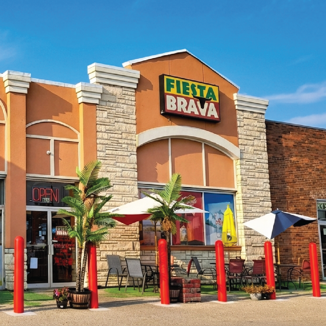 Exterior of Fiesta Brava restaurant with palm plants, outdoor seating, umbrellas, and red bollards under a clear blue sky.