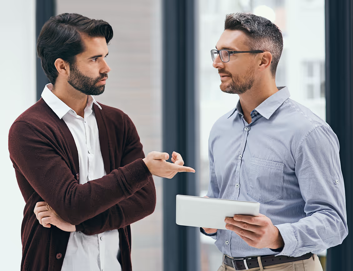Two men having a serious conversation in a modern office, one holding a tablet and the other gesturing with his hand.