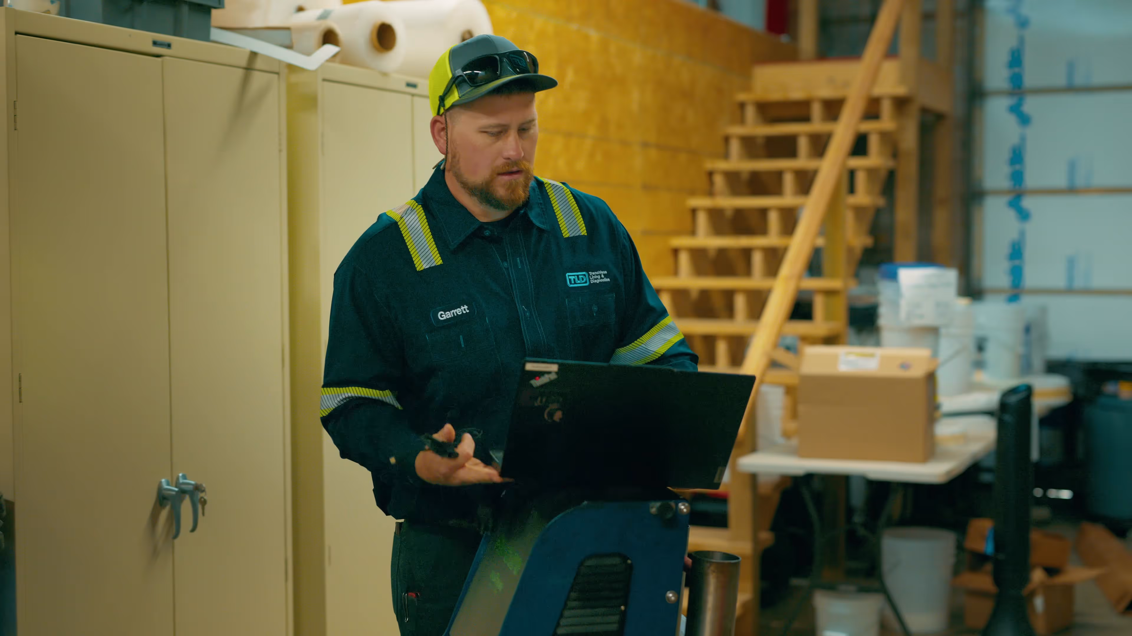 Man wearing a high-visibility work shirt and cap standing indoors, looking at a laptop on a stand in a workshop with storage cabinets and a wooden staircase in the background.