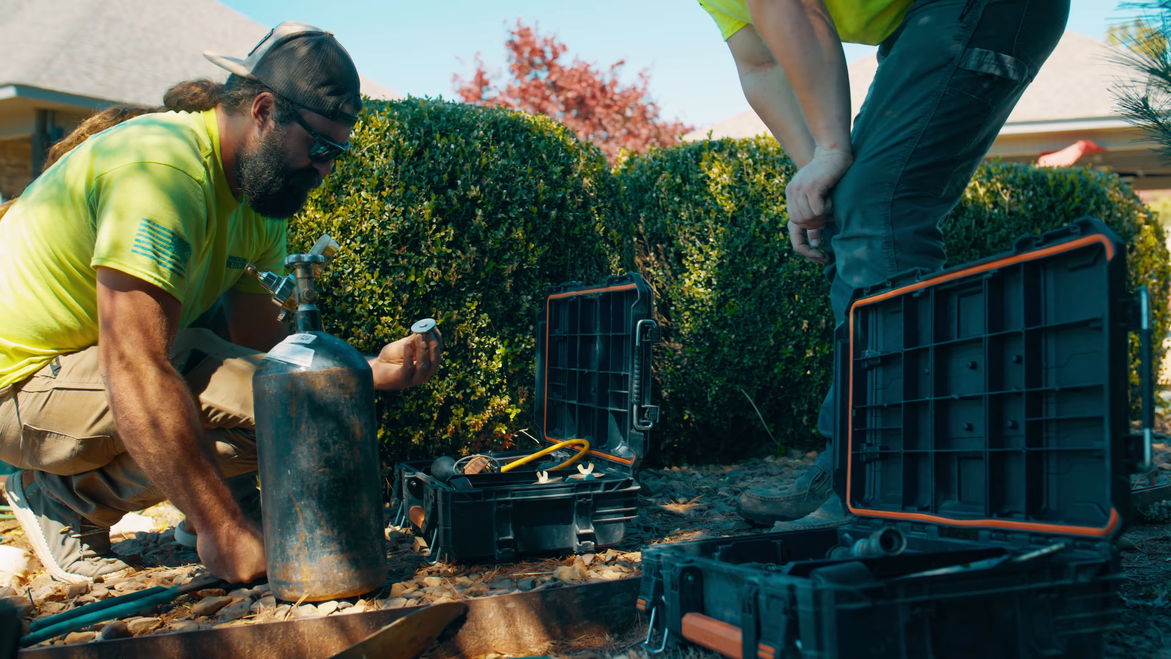 Two workers in neon yellow shirts working outdoors with toolboxes and a gas cylinder near green shrubs.