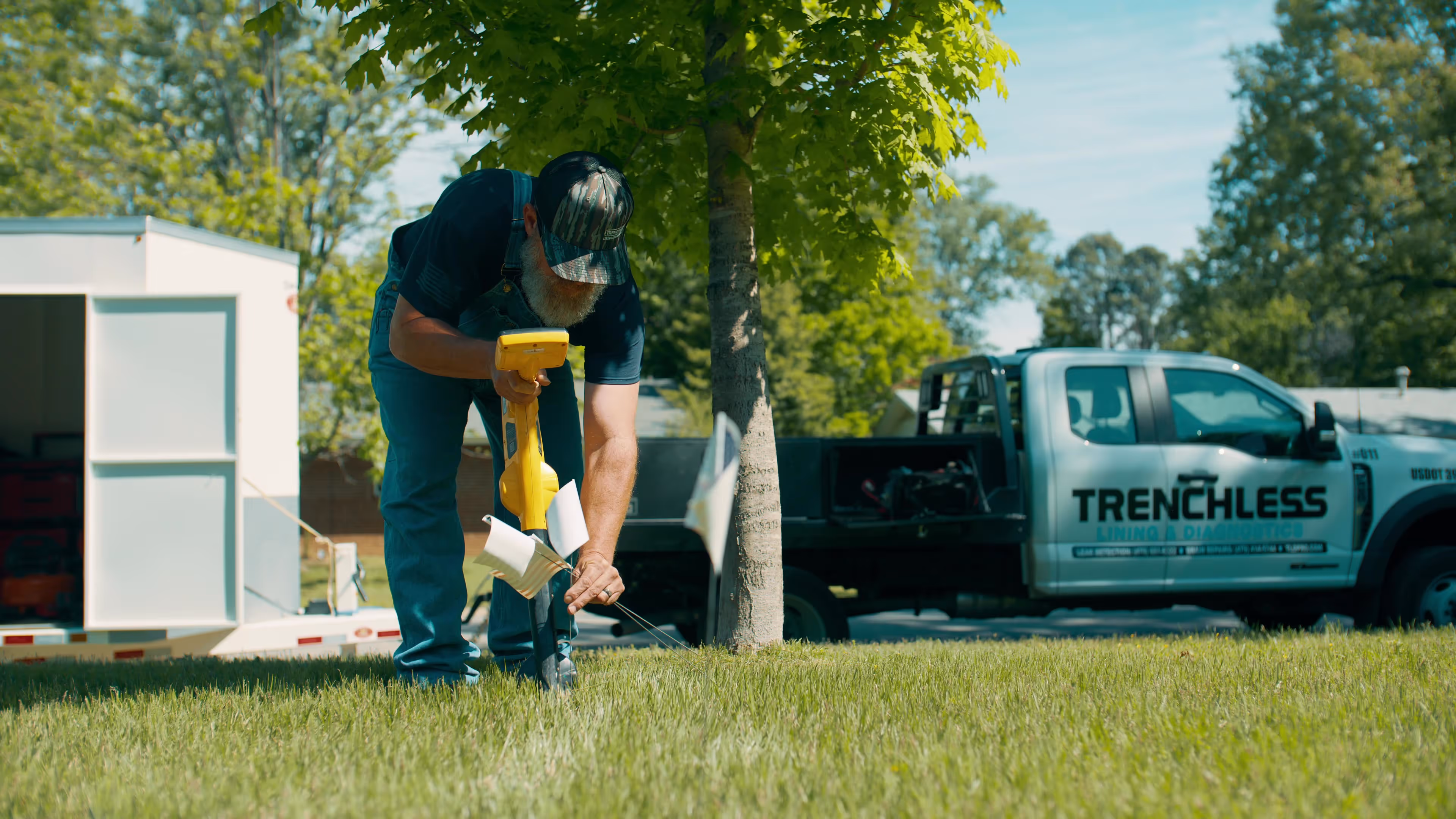 Man in overalls using a yellow ground-penetrating radar tool in a grassy area near a tree with a truck labeled 'Trenchless' in the background.
