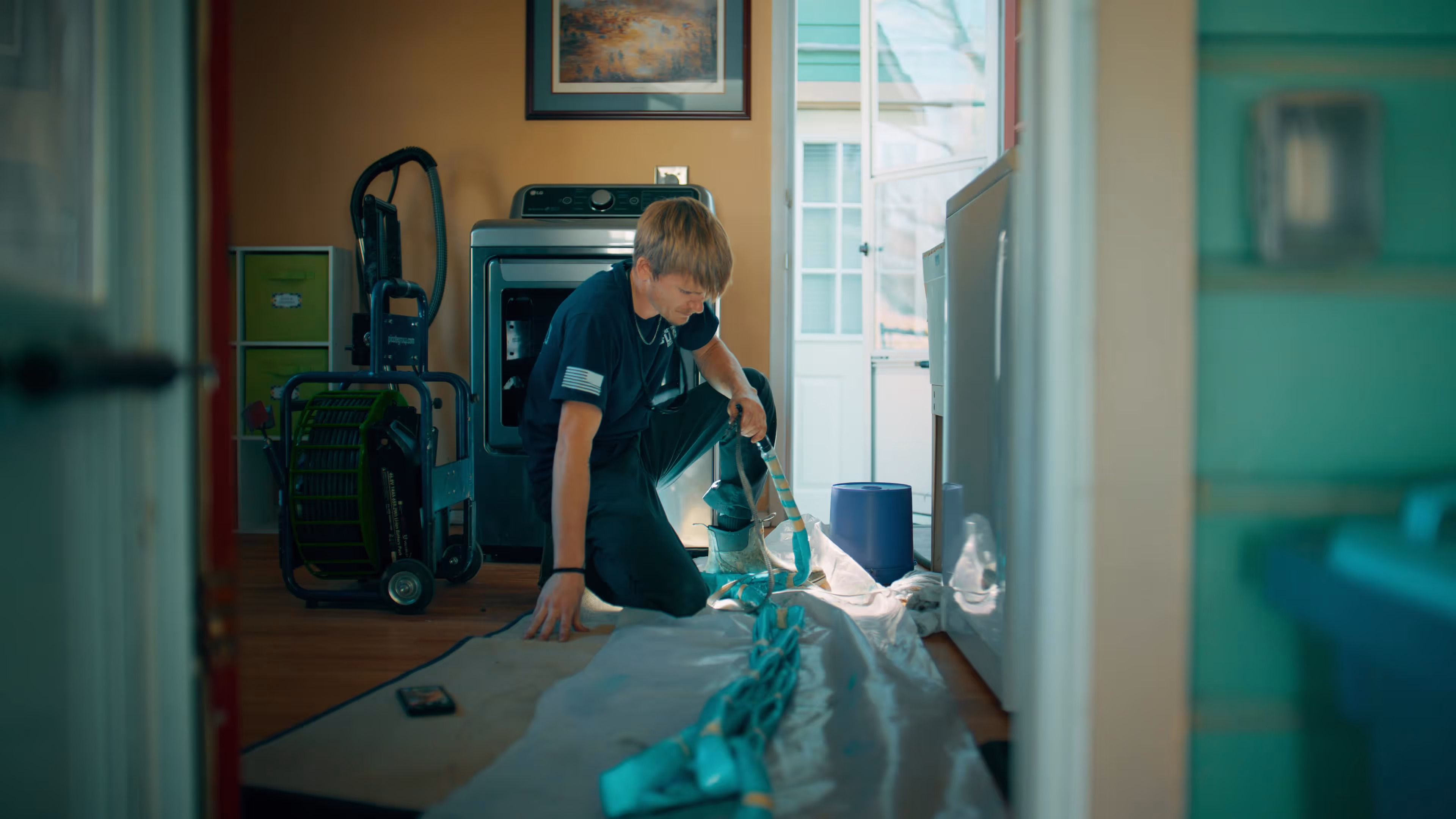 Man kneeling on the floor organizing blue NuDrain pipes in a laundry room with a washing machine and door in the background.