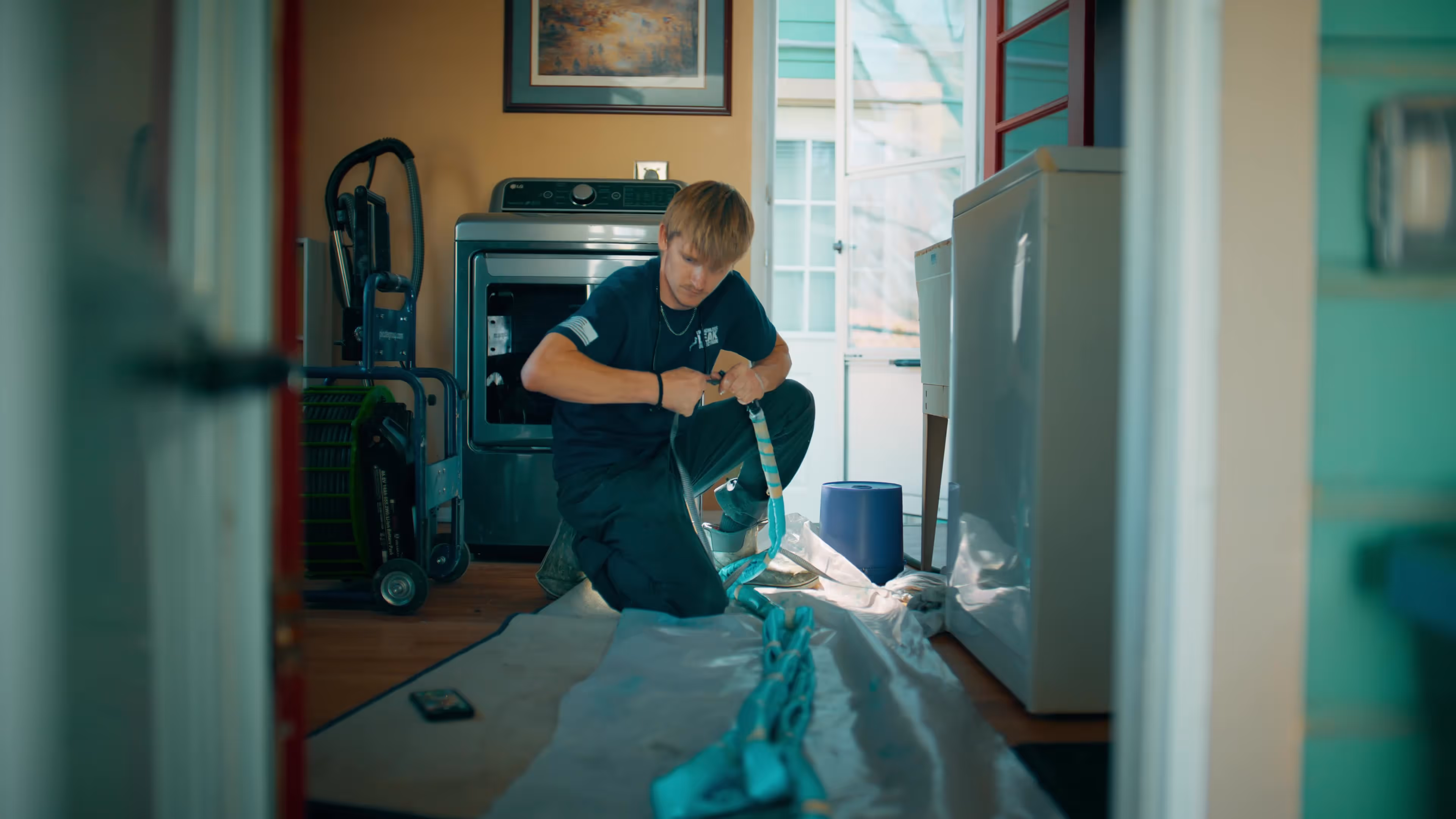 Technician kneeling on the floor working on a blue  NuDrain pipe inside a laundry room with a washer and dryer.
