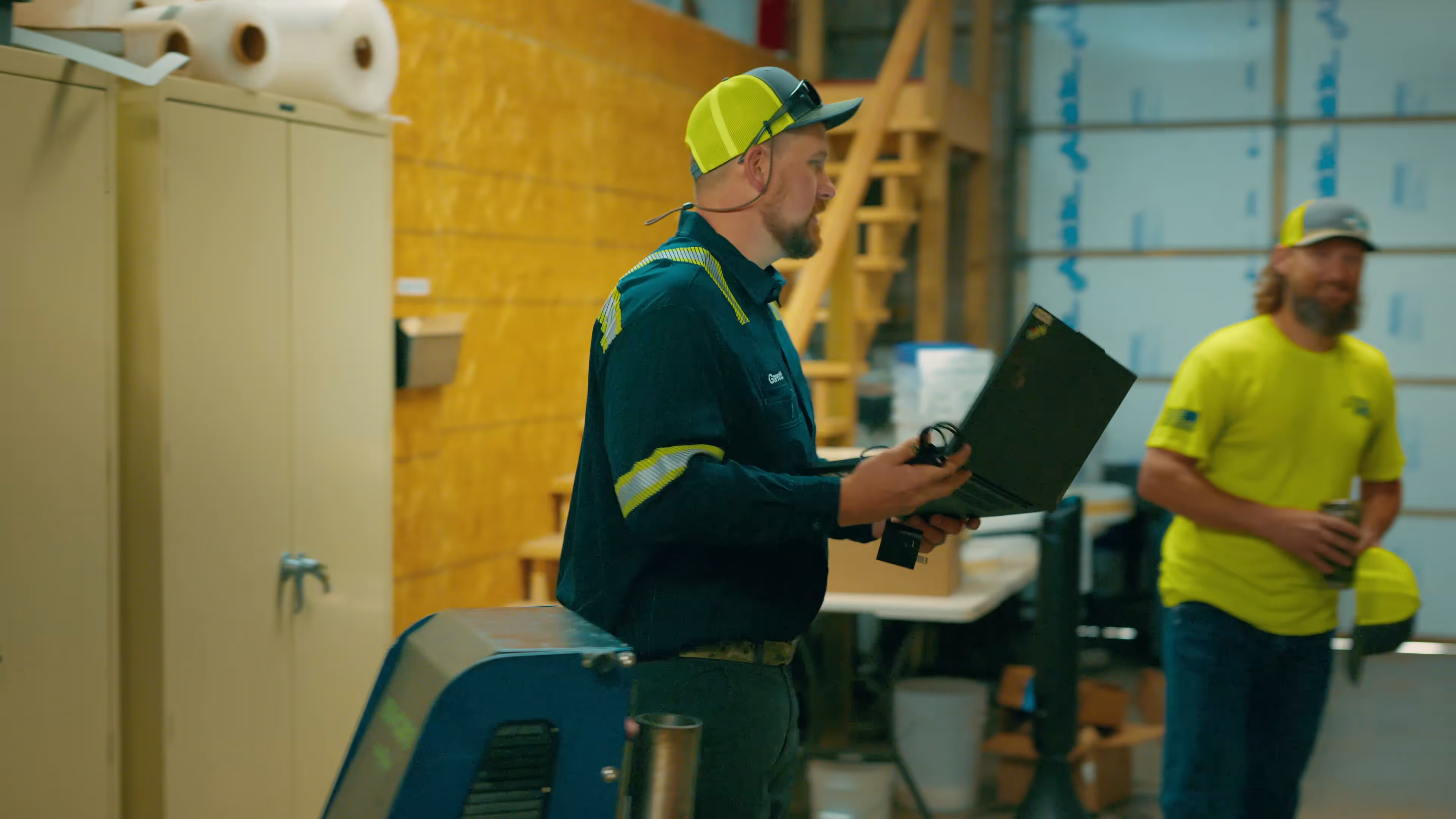 Two workers in a workshop; one wearing a yellow and black cap and holding a laptop, the other in a bright yellow shirt holding a drink and a yellow helmet.