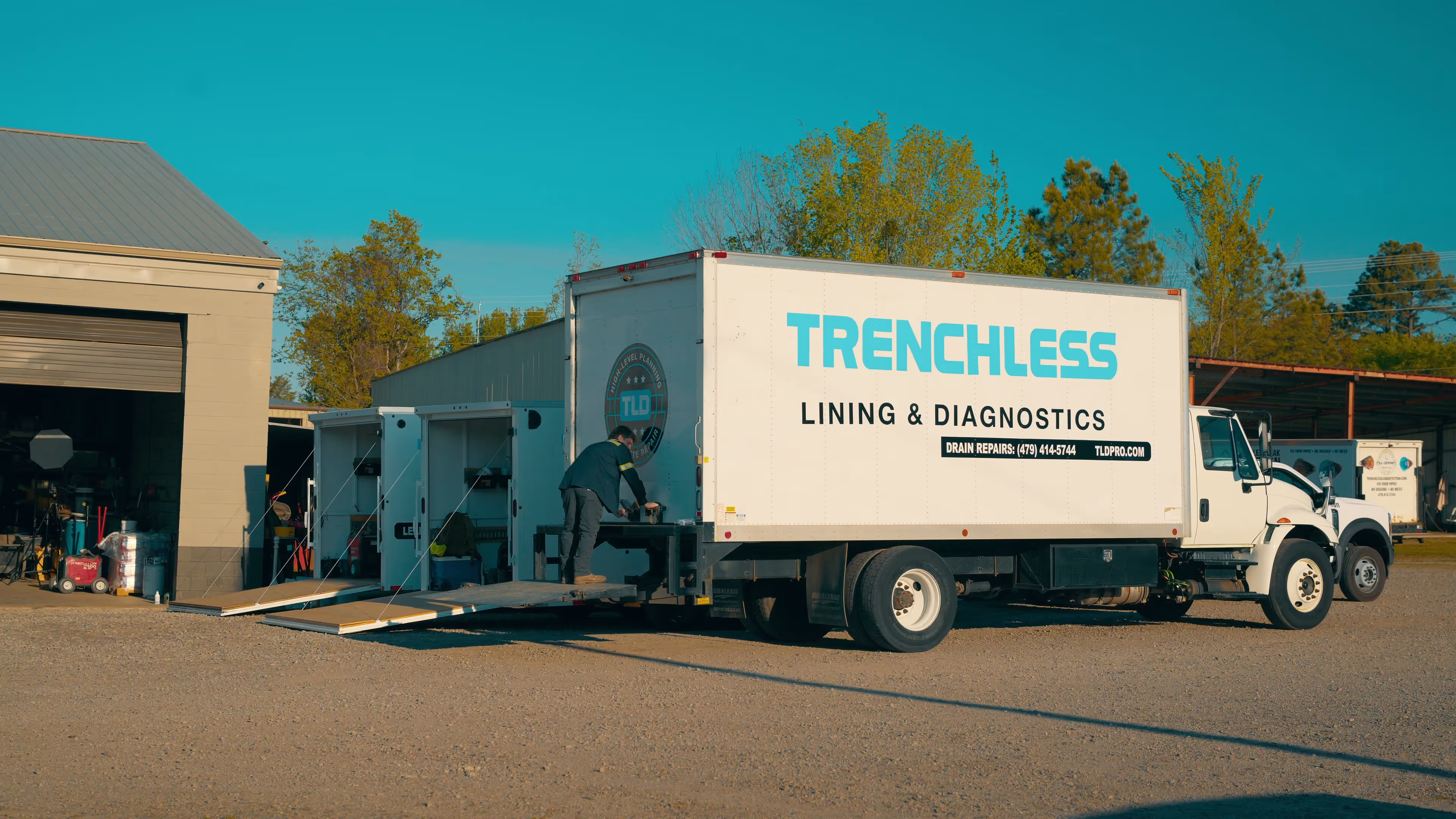 A worker stands on the back ramp of a white truck labeled 'Trenchless Lining & Diagnostics' parked outside a garage.