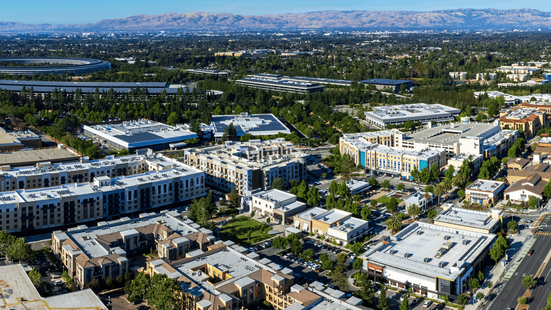 Aerial view of a modern urban area with multiple apartment buildings, offices, green spaces, and distant mountains under a clear blue sky.
