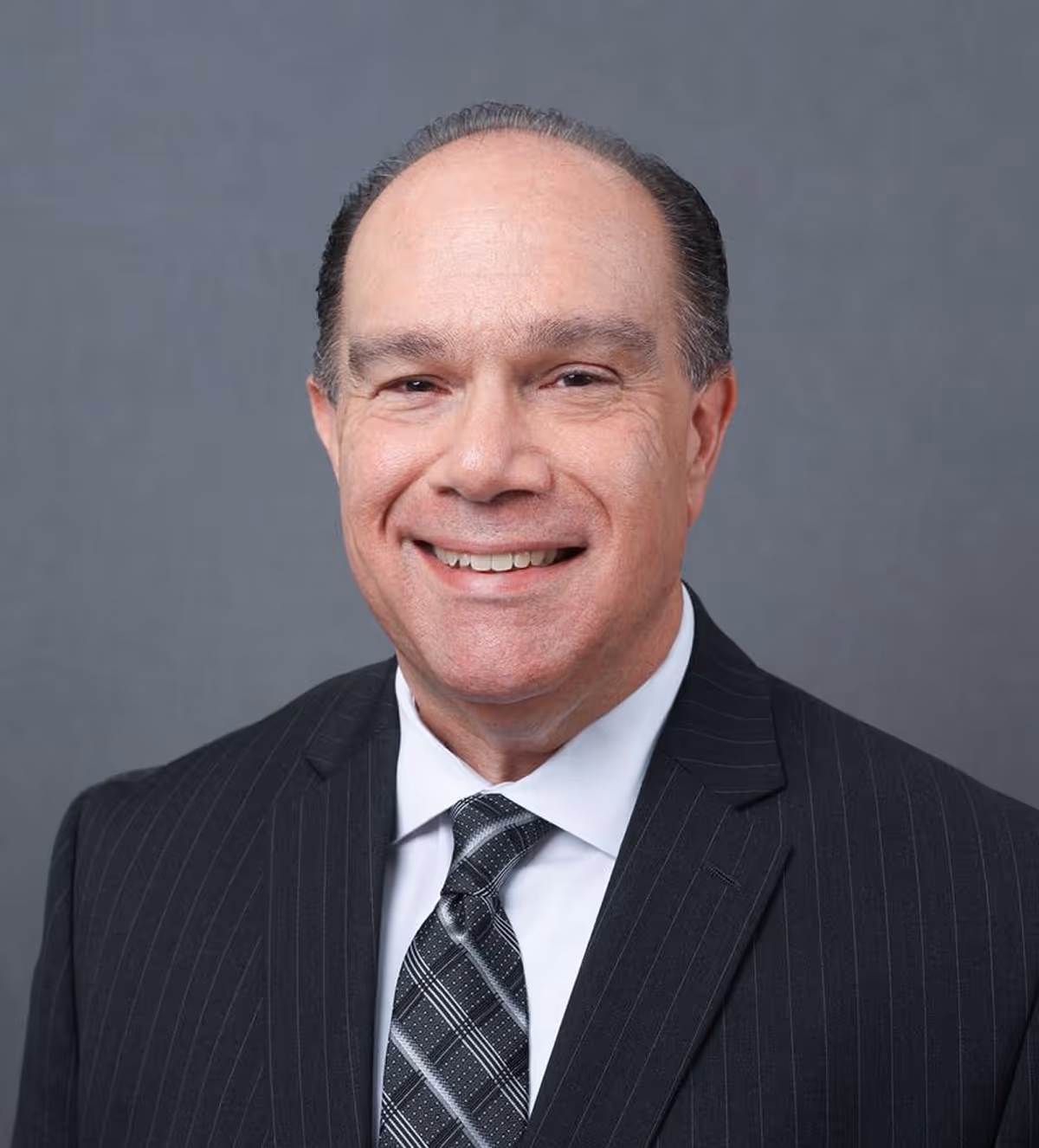 Smiling middle-aged man in a black pinstripe suit with a white shirt and patterned tie against a gray background.