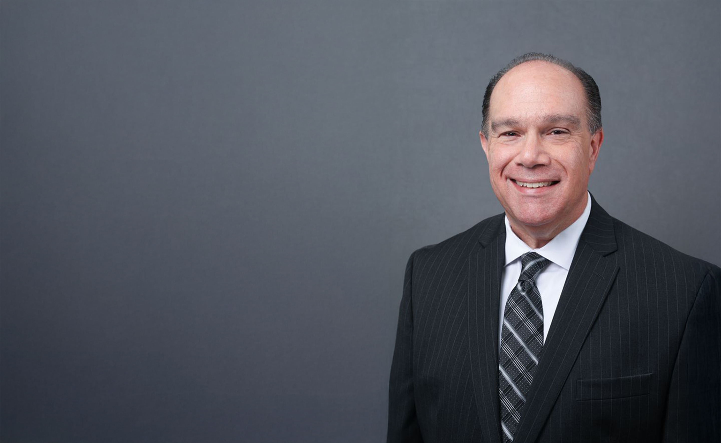 Smiling middle-aged man in a dark pinstripe suit and patterned tie against a plain gray background.