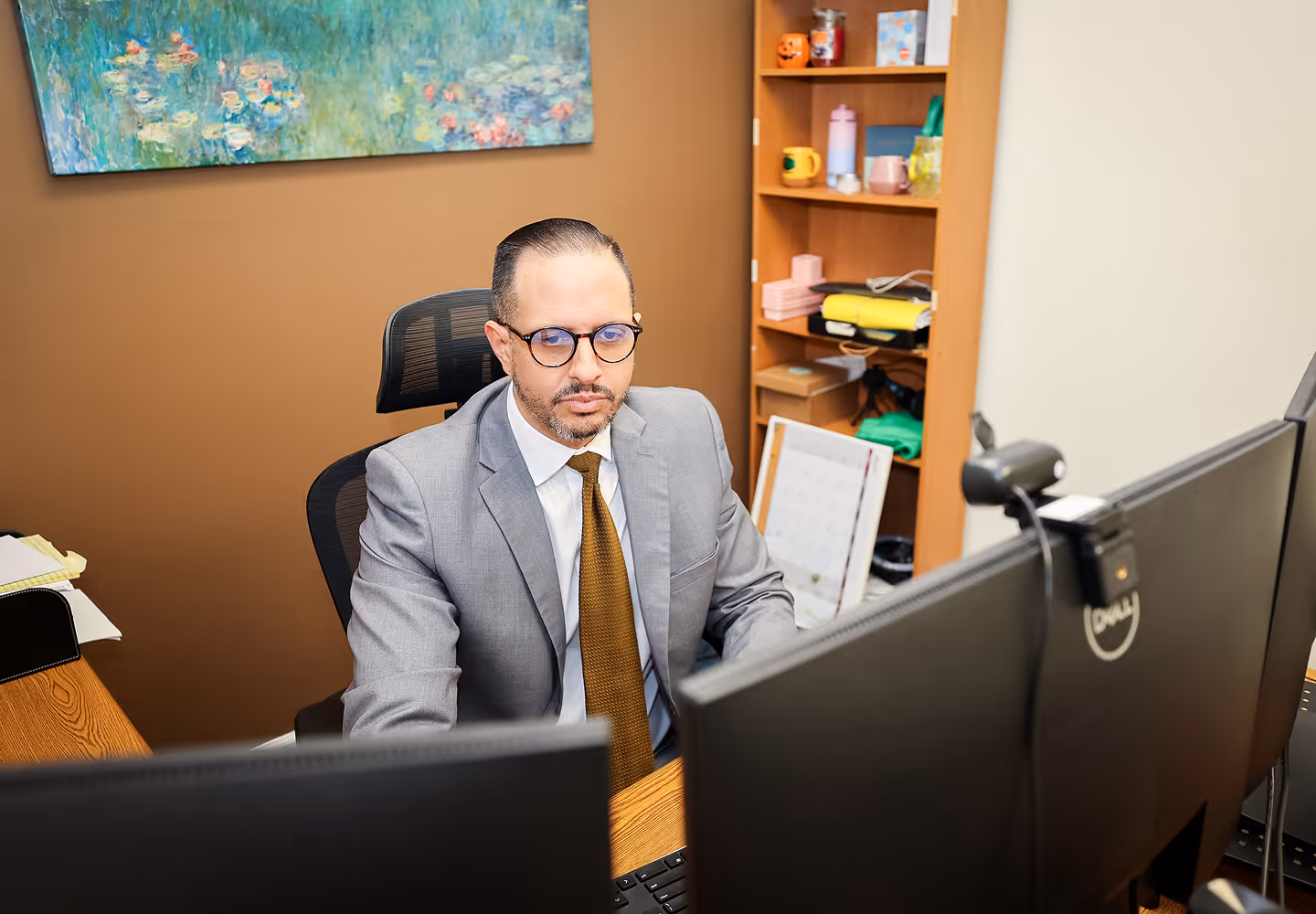 Man in gray suit and glasses working at a desk with two Dell monitors in an office.