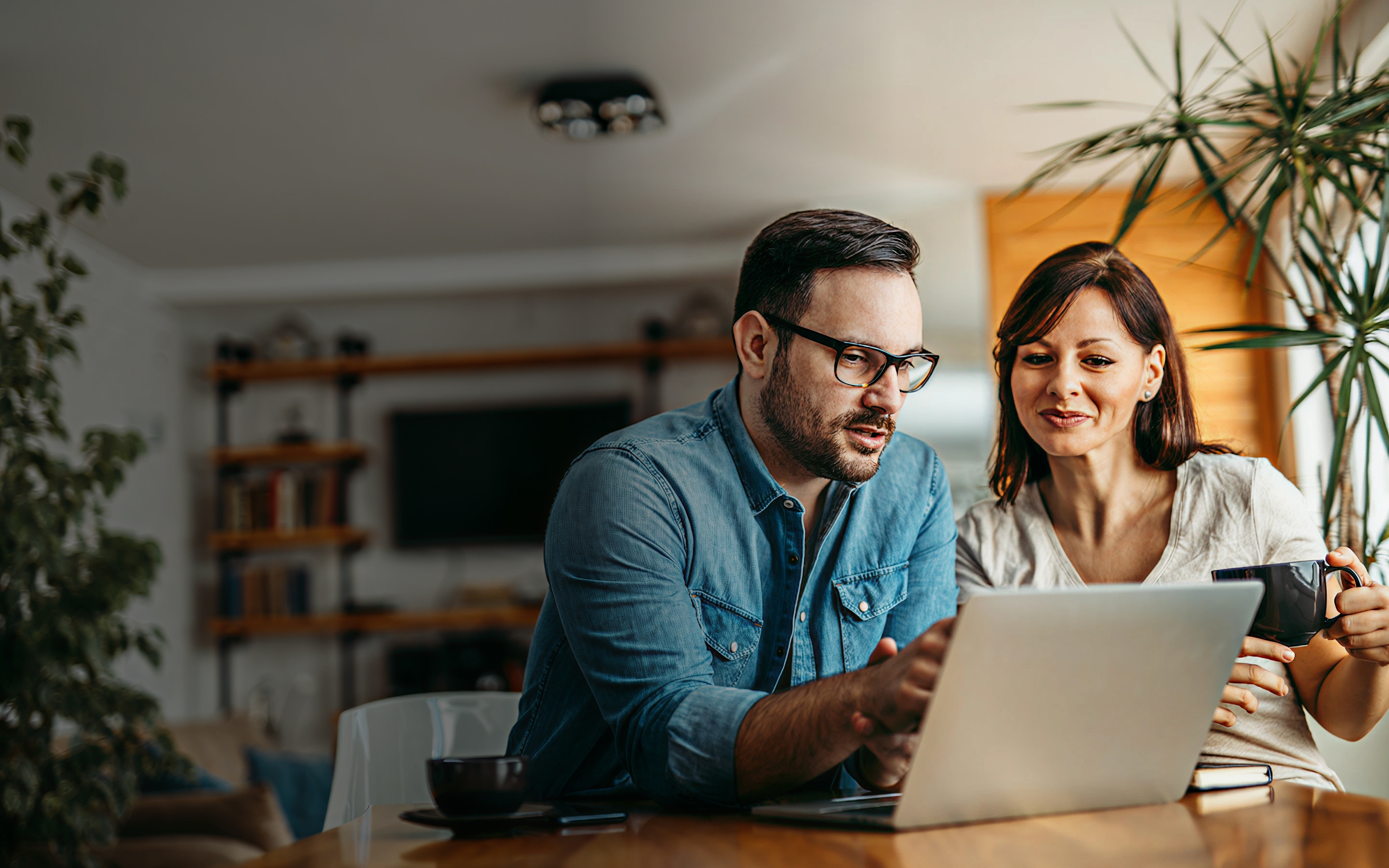 Man and woman sitting at a table looking at a laptop while the woman holds a mug.