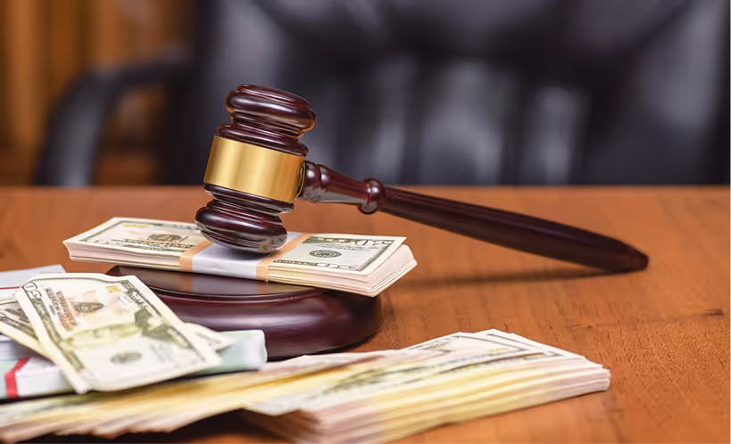 Wooden gavel resting on a base with stacks of US dollar bills on a wooden table.