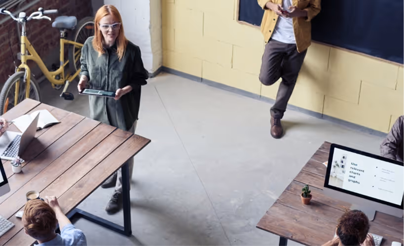 Top-down view of a modern office with people working at desks with laptops and a woman standing holding a tablet, a yellow bicycle against a brick wall.