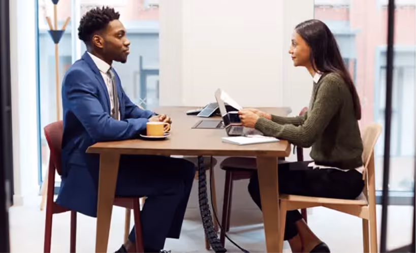 Two professionals sitting across from each other at a table, engaged in a discussion with a laptop and documents.