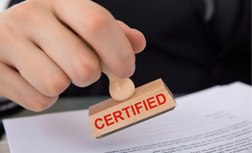 Hand holding a wooden stamp with the word CERTIFIED in red, pressing it onto a document.