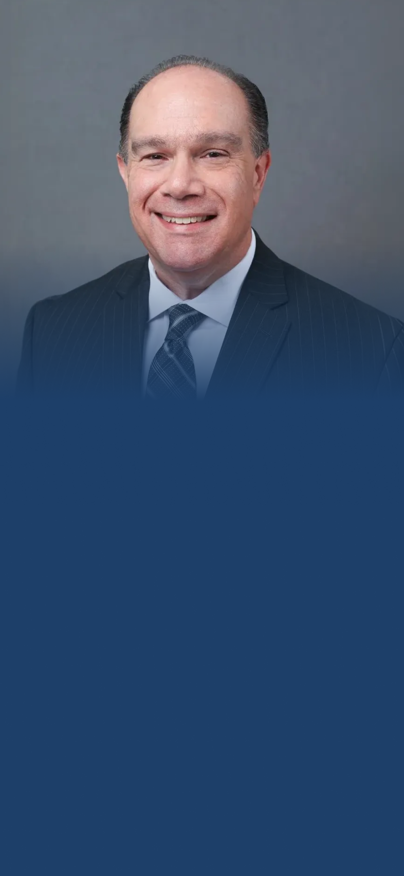 Smiling middle-aged man wearing a dark pinstripe suit, white shirt, and patterned tie against a plain gray background.