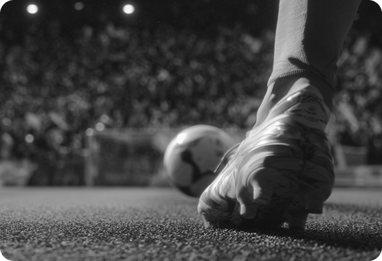 Close-up of a soccer player's cleated foot near a soccer ball on a field with a blurred stadium crowd in the background.