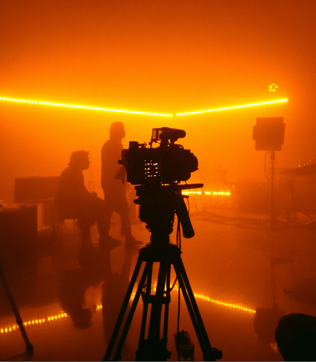 Silhouettes of two people and a video camera on a tripod in a dim, orange-lit studio with LED strip lights.