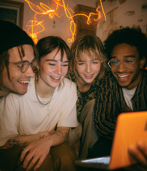 Four friends smiling and looking at a laptop screen with warm ambient lighting in the background.