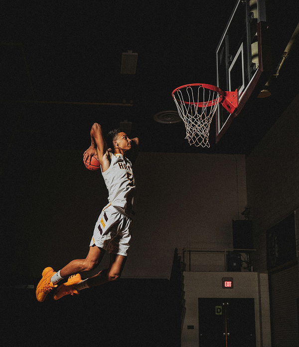 Basketball player mid-air approaching the rim for a dunk in a dimly lit indoor court.