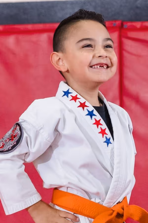 A cute little kid wearing a white gi and orange belt is standing in front position waiting for his karate class to start.