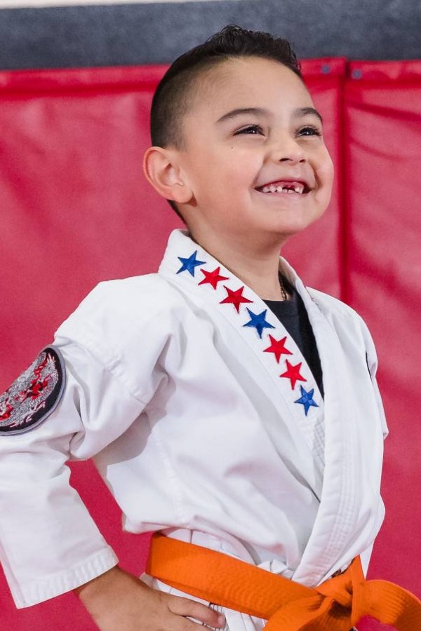 A cute little kid wearing a white gi and orange belt is standing in front position waiting for his karate class to start.