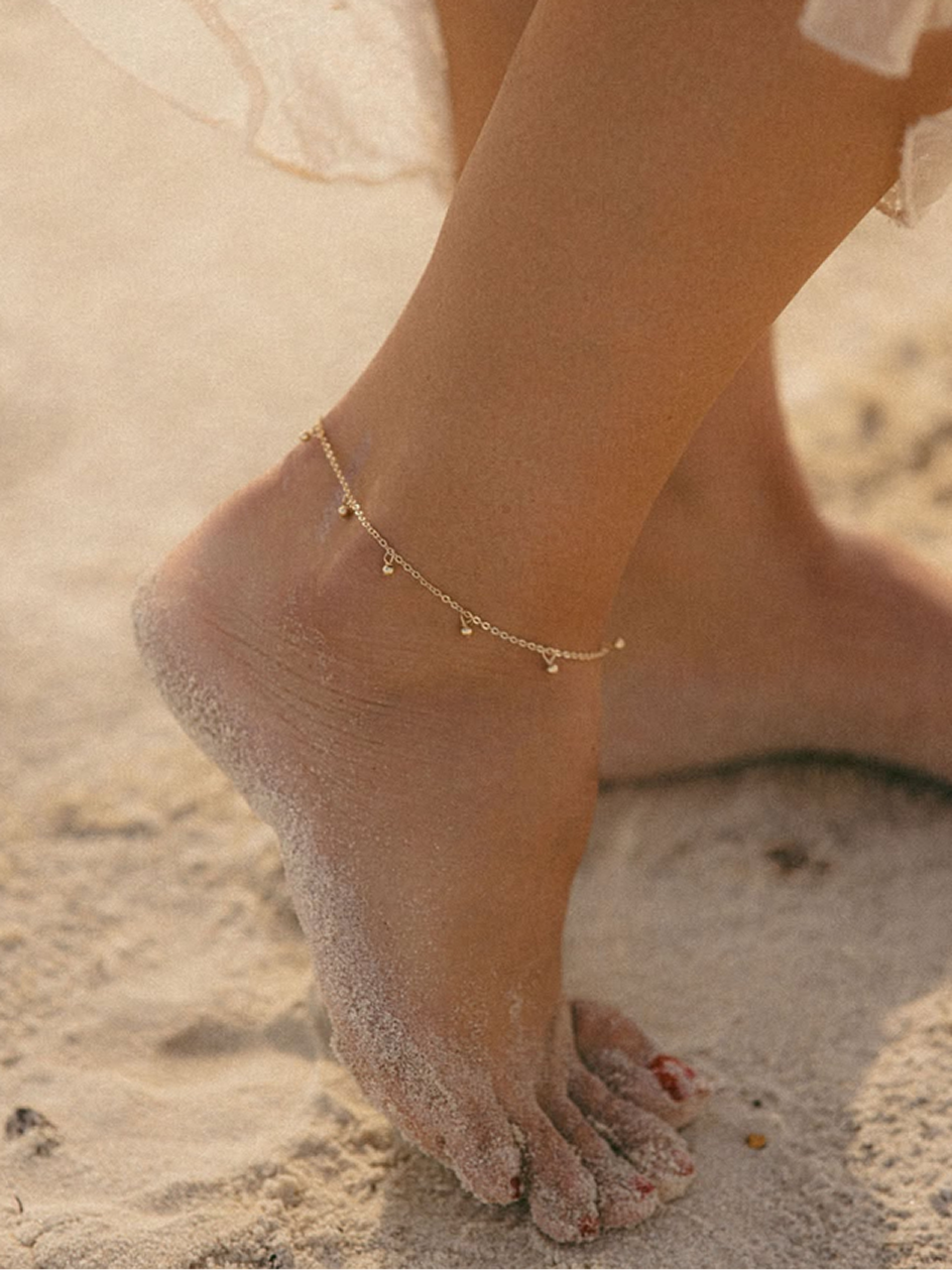 Close-up of a sandy foot wearing a delicate gold anklet on a beach.