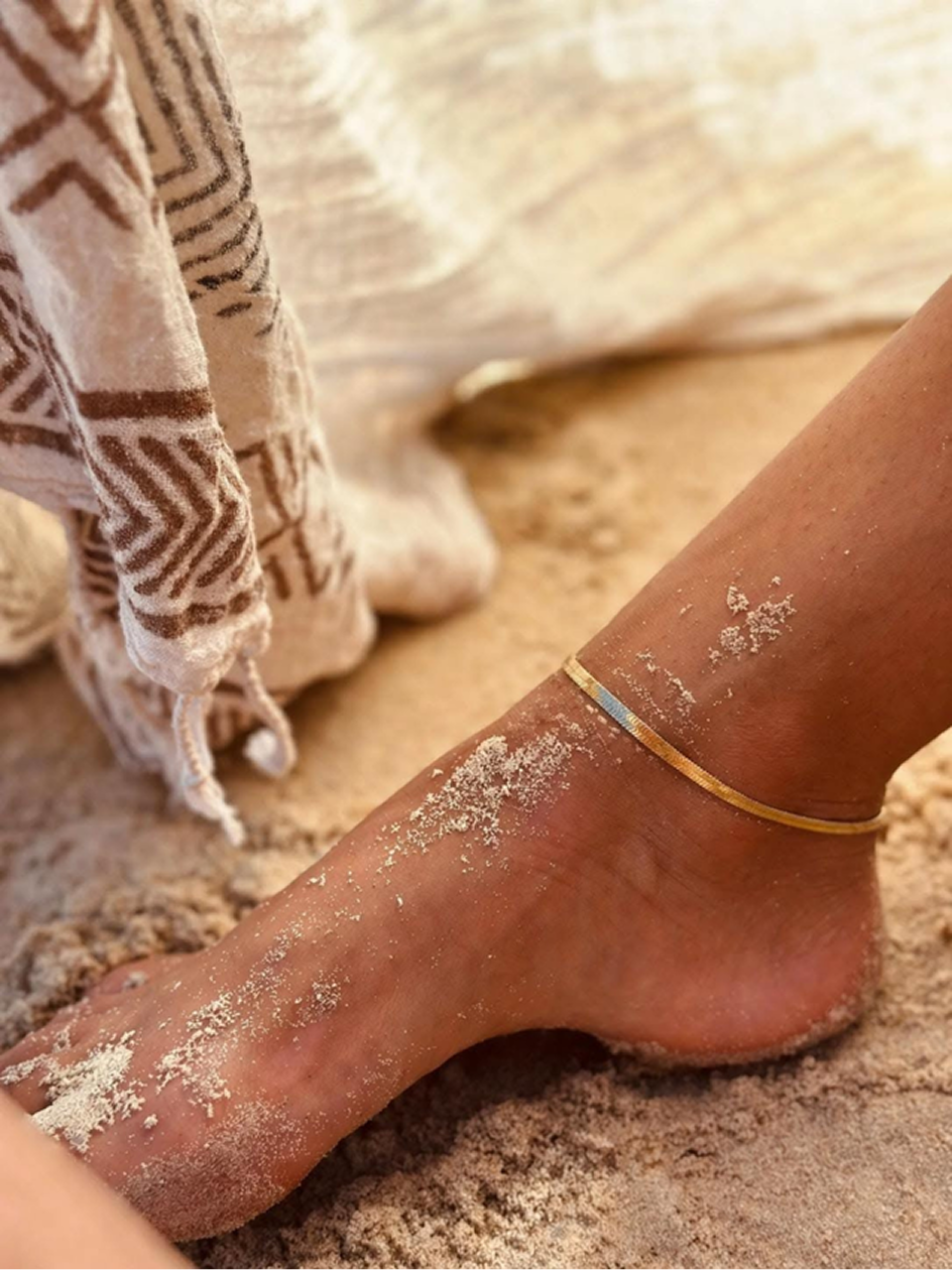 Sandy foot on beach wearing a thin gold anklet beside a patterned beige cloth.