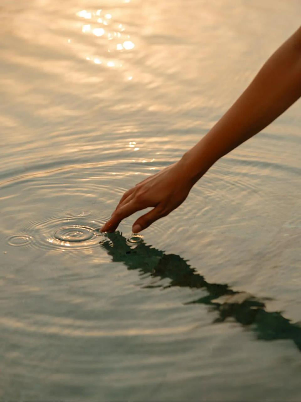 Hand touching calm water surface creating gentle ripples with sunlight reflections.