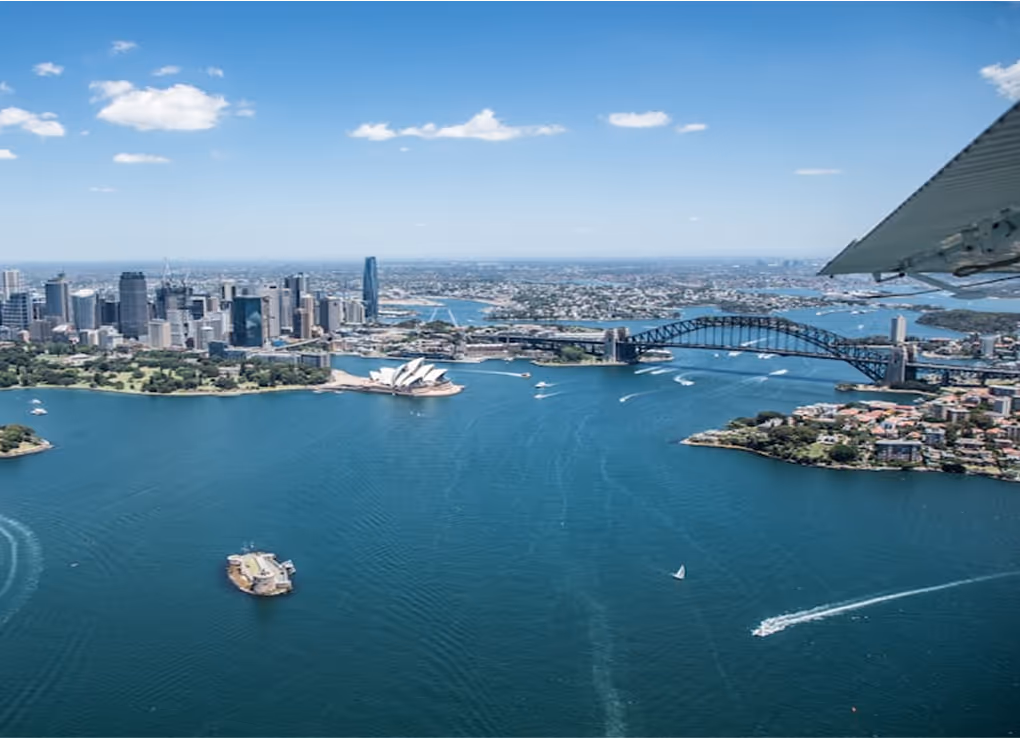 Aerial view of Sydney Harbor with the Sydney Opera House and Sydney Harbour Bridge under a bright blue sky.