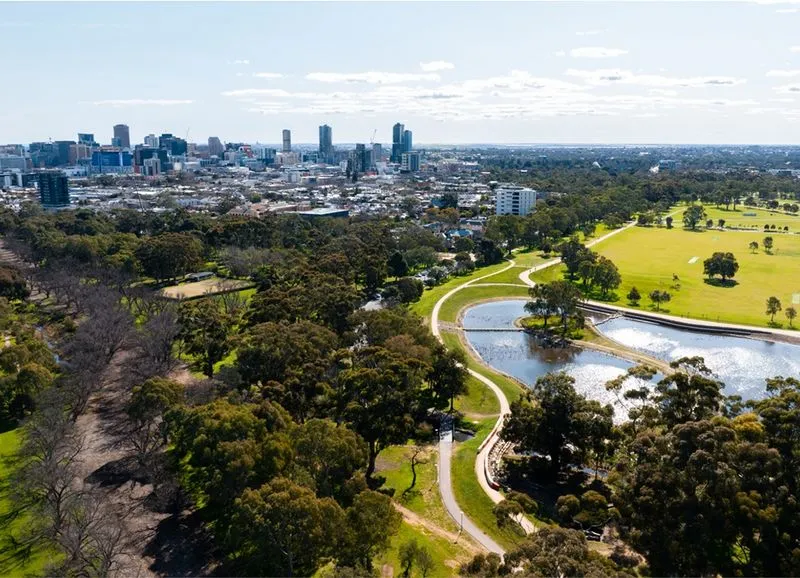 Aerial view of a cityscape with a large park featuring walking paths, trees, and ponds in the foreground.
