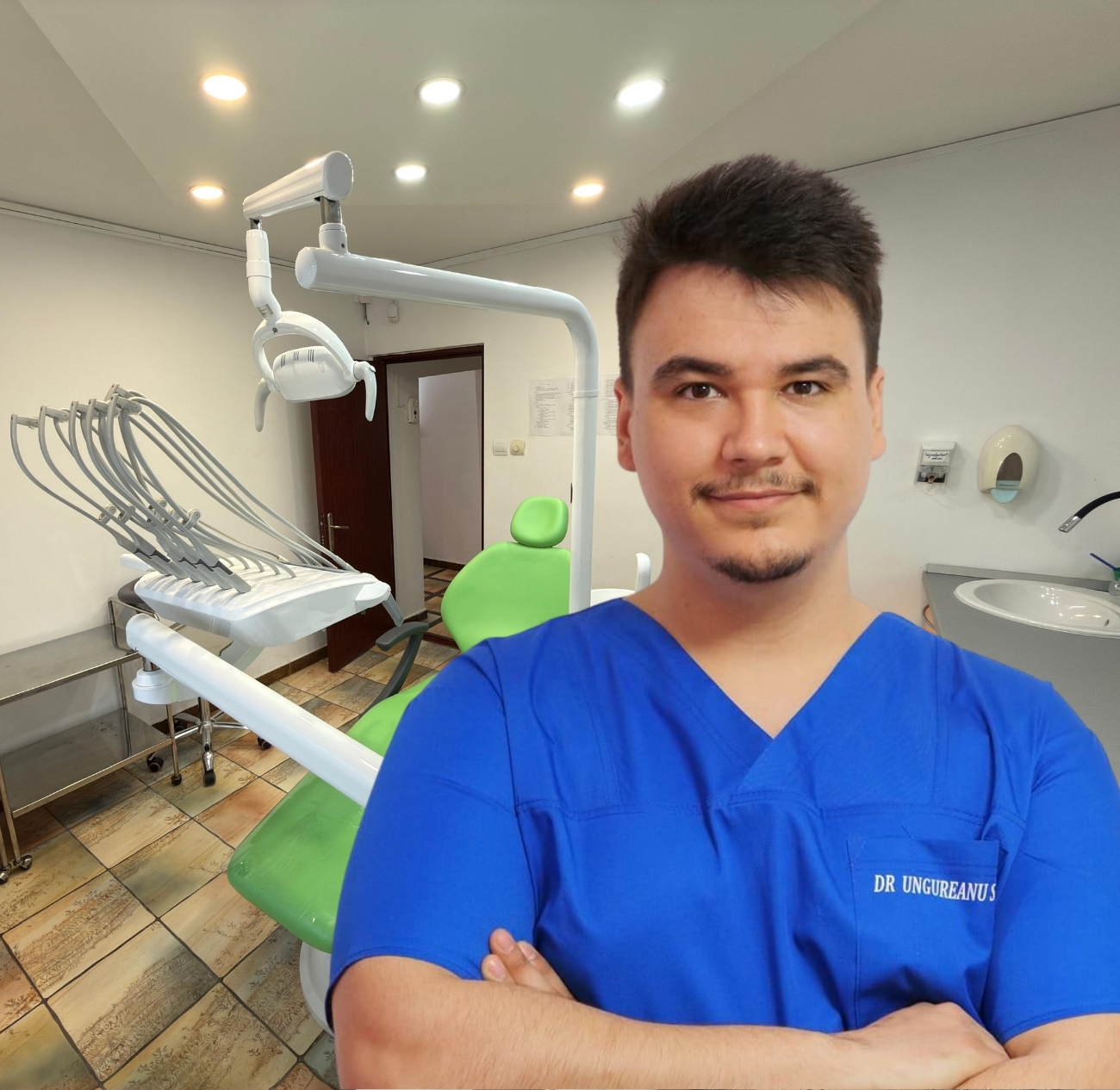 Medic dentist tânăr îmbrăcat în halat albastru, cu brațele încrucișate, în cabinet stomatologic cu scaun verde și echipamente dentare albe.