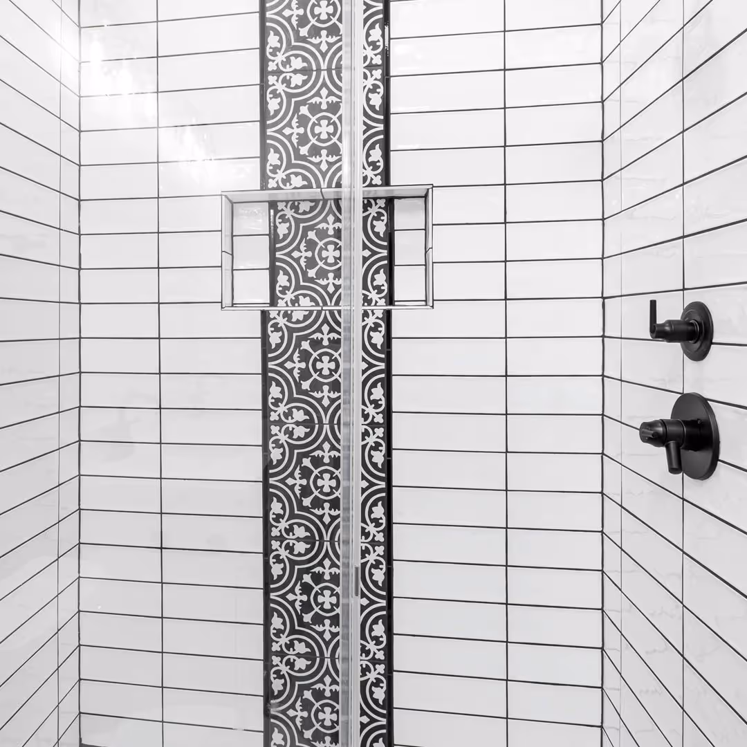Shower interior with white rectangular tiles, a vertical black and white patterned tile strip, built-in shelf, and black faucet controls.