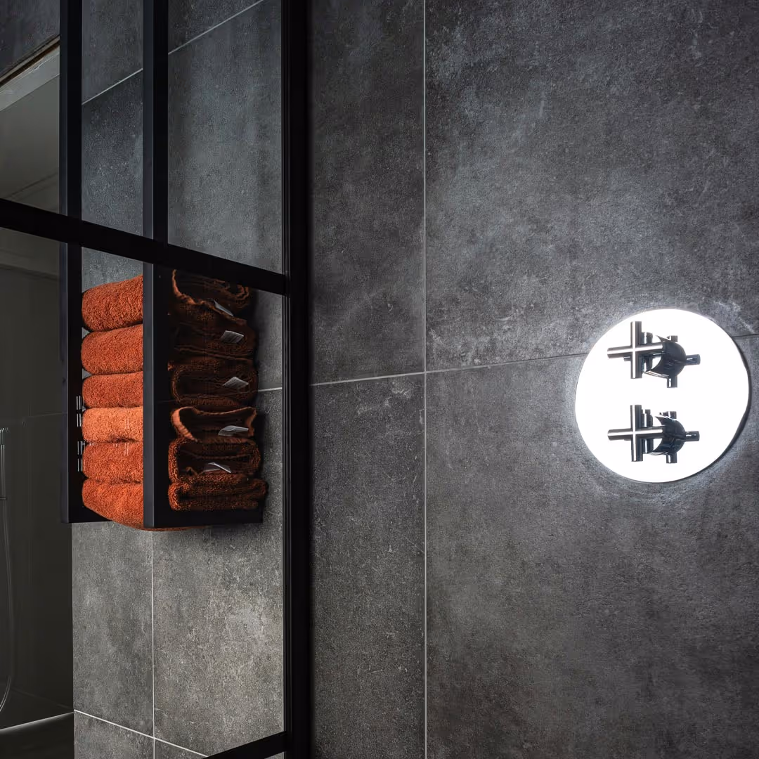 Wall-mounted shelves with neatly folded orange towels on dark grey tiled wall beside round chrome shower control in modern bathroom design.