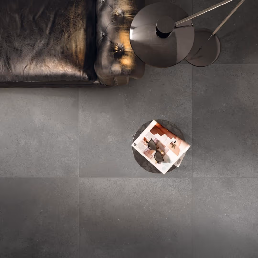 Top-down view of dark gray textured floor tiles next to a dark leather sofa and a small round table holding a magazine and sunglasses.