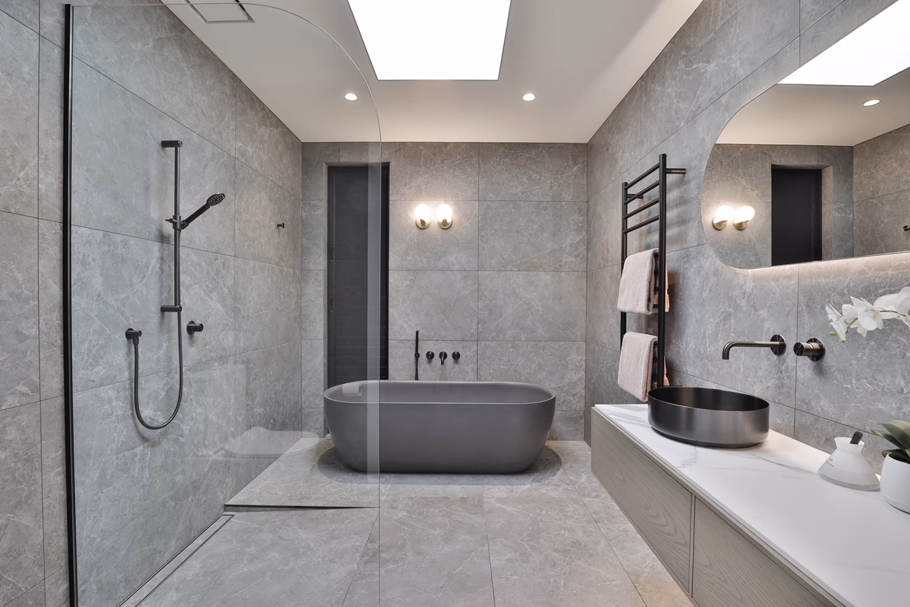 Modern bathroom with grey stone tiles, freestanding grey bathtub, glass shower enclosure, black oval sink, and towel rack with pink towels in contemporary bathroom design.