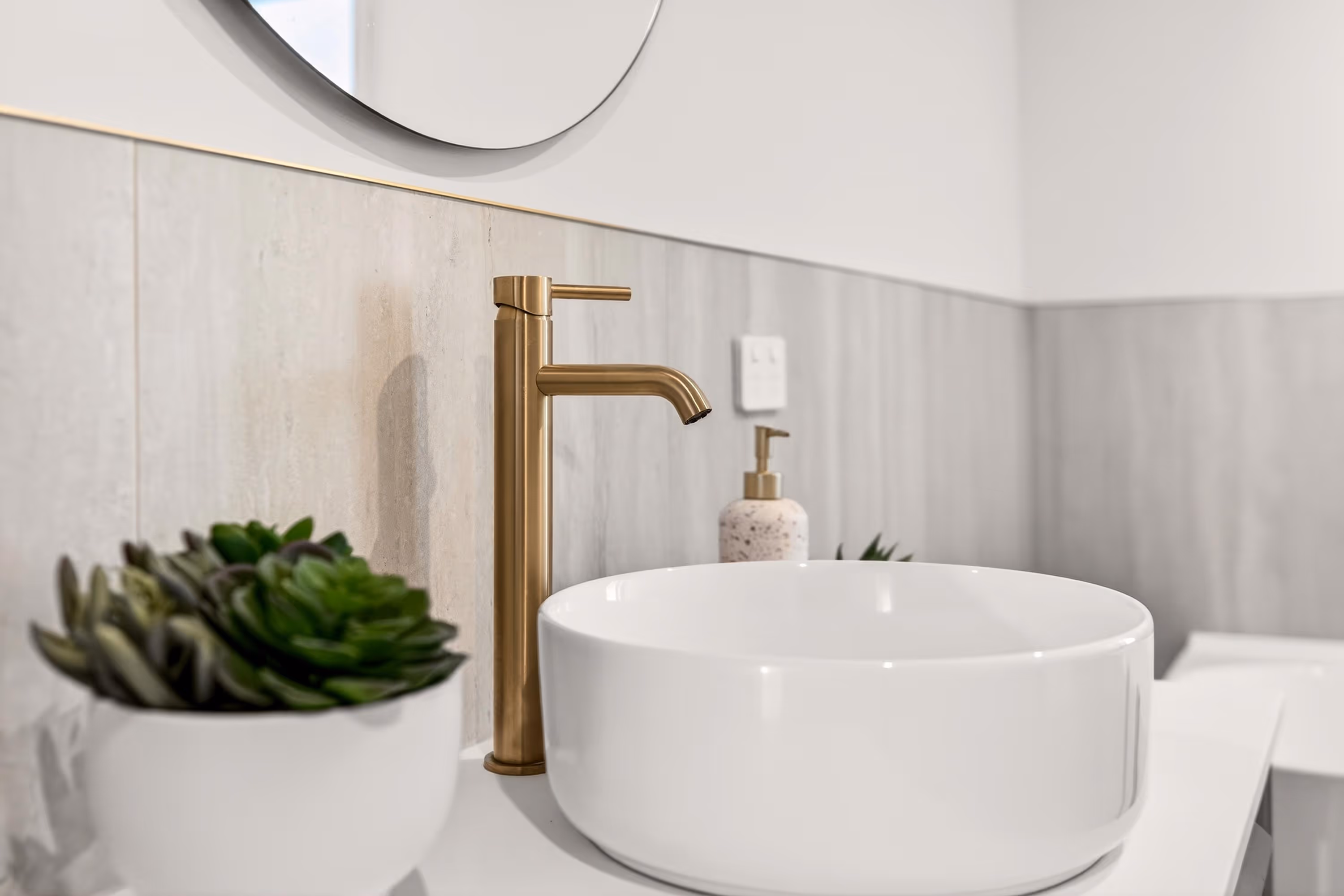 Modern bathroom sink with matte gold faucet, round white basin, wall tiles, and small potted succulent plant in contemporary bathroom design.