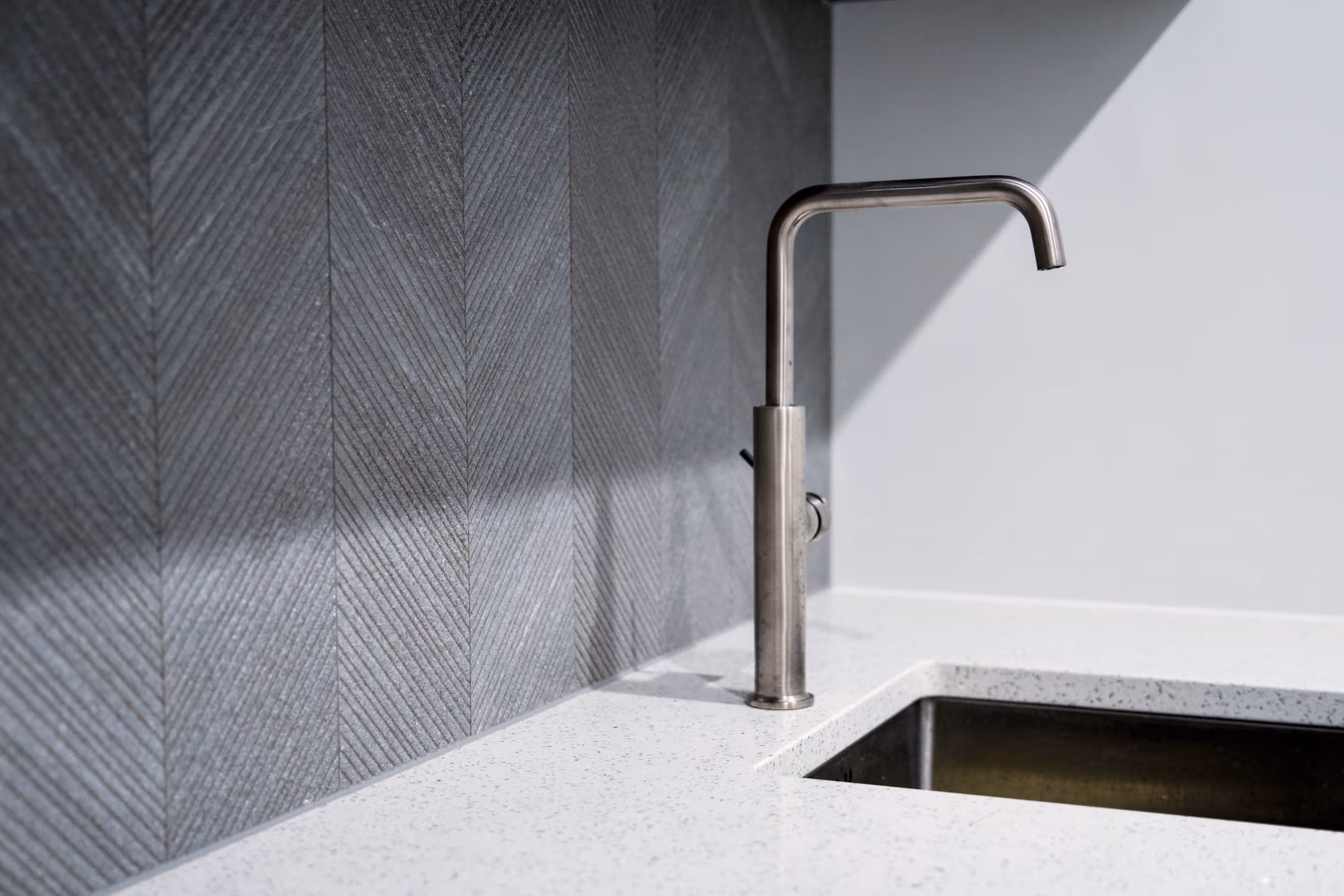 Modern stainless steel kitchen faucet over white speckled countertop with dark geometric tile backsplash in contemporary kitchen design.